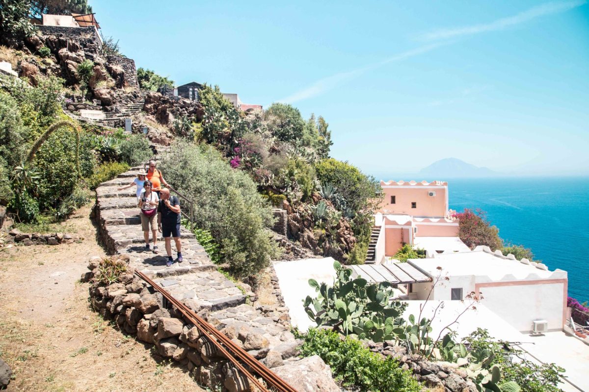 Gruppo di escursionisti che cammina su un sentiero in pietra tra la vegetazione mediterranea delle Isole Eolie, con case bianche e rosa sullo sfondo e il mare azzurro all’orizzonte.