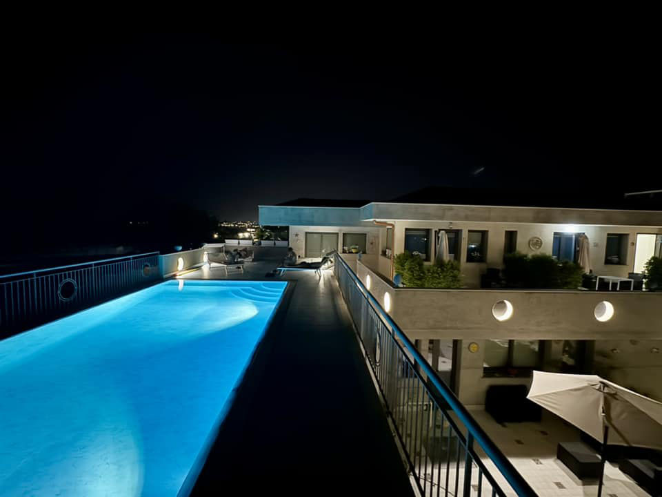 Vista di un resort moderno con piscina panoramica e area relax su una terrazza sopraelevata, circondato da vegetazione e case circostanti