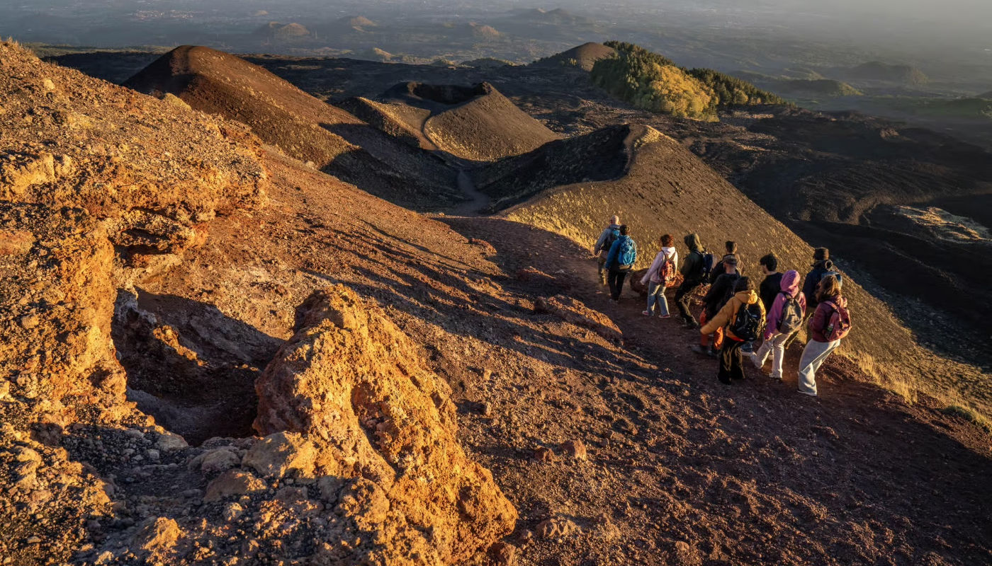 Escursionisti che camminano in fila lungo un sentiero di rocce vulcaniche rosse sull’Etna al tramonto, con crateri e colate laviche sullo sfondo.