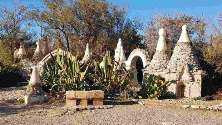Mini villaggio di trulli in pietra con tetti conici a Barcarello, Sicilia, circondato da piante mediterranee come agavi e fichi d’india, sotto un cielo azzurro limpido.